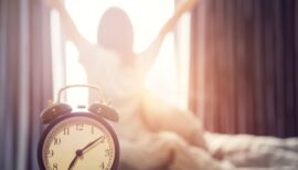 Closeup alarm clock having a good day with background happy woman stretching in bed after waking up, sunlight in morning.